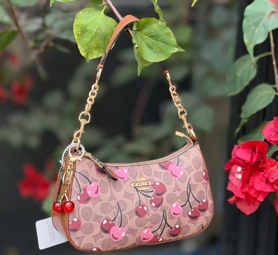 compact, crescent-shaped handbag crafted in Coach's signature logo canvas with leather trim. It features top zip closure, gold chain-link shoulder straps, and a playful 3D cherry charm.